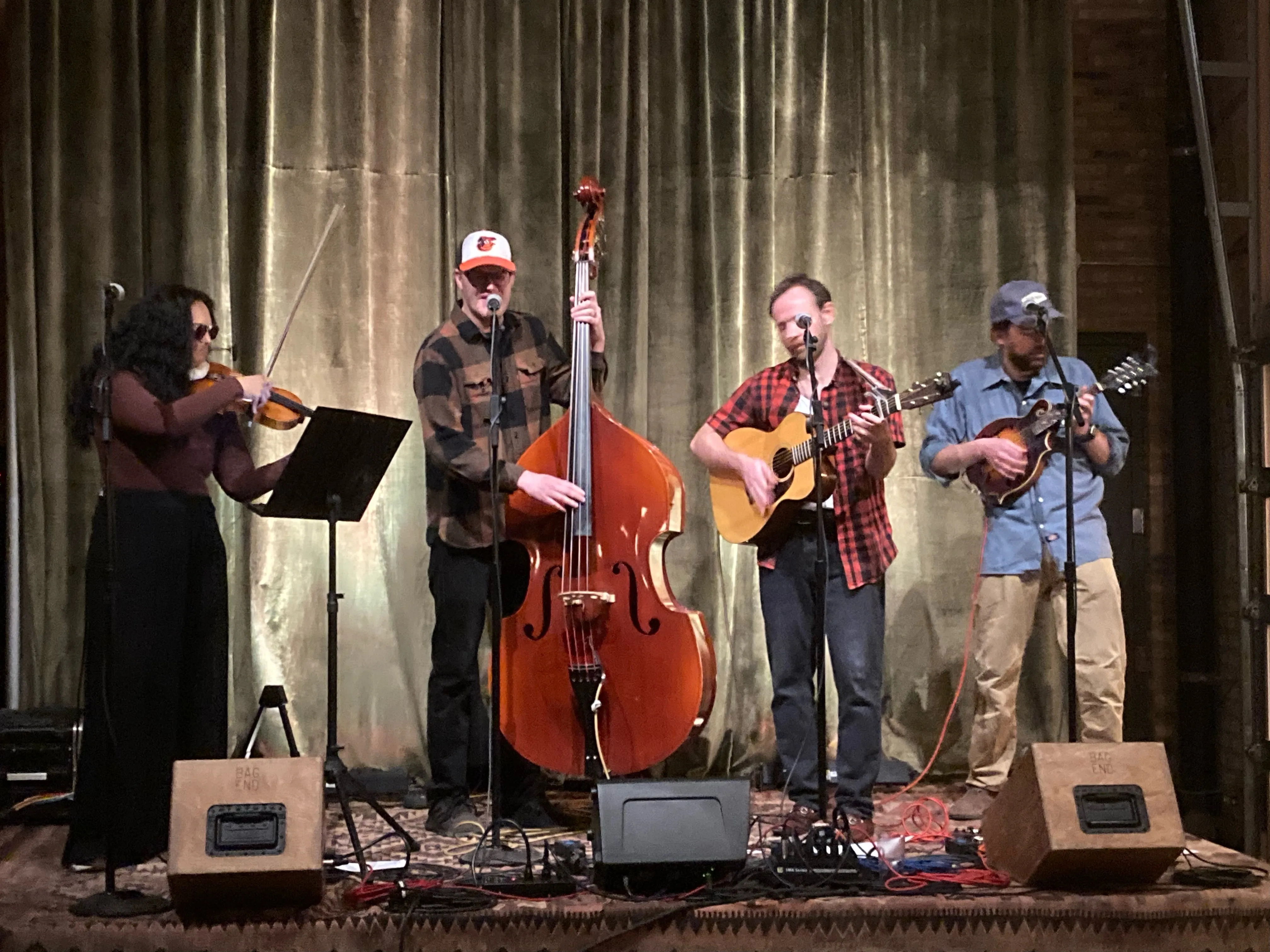 Potomac Bombs on stage in front of a green velvet curtain, playing fiddle, upright bass, guitar and mandolin.